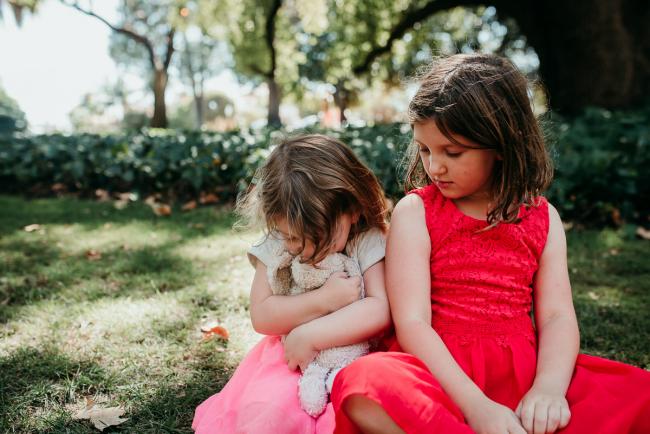 Family-Photographer-Perth-Hyde-Park-33-of-34 Little girl cuddling her bunny as she leans against her big sister who is looking down at her during a Perth family photography session at Hyde Park