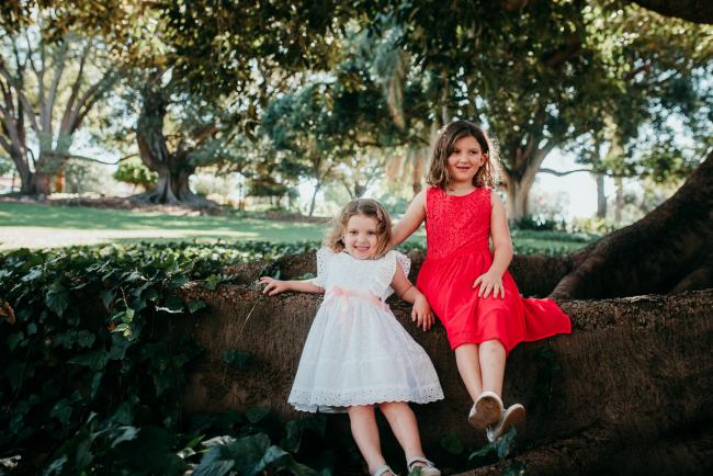 Family-Photographer-Perth-Hyde-Park-7-of-34 Girl in a red dress sitting on a tree as her little sister in a white dress leans against the tree during a Perth family photography session at Hyde Park