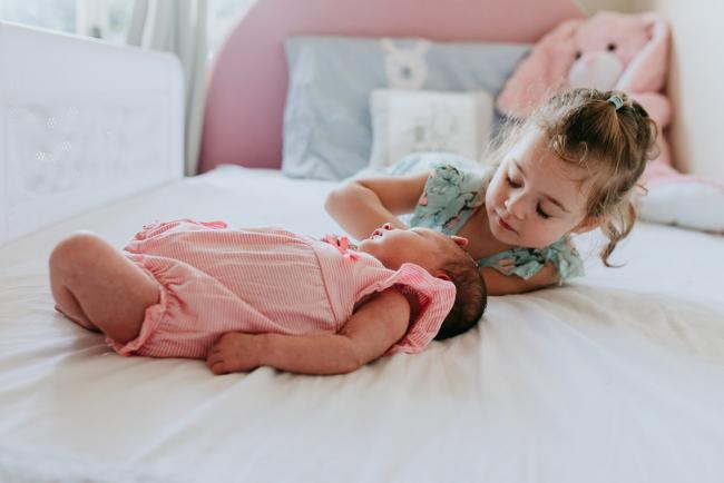 New baby laying on a bed as her big sister lays near her and touches her head during a Perth newborn lifestyle photography session