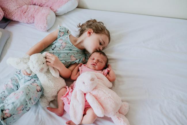 New baby and older sister lay on a bed with their bunnies as big sister kisses new baby during a Perth newborn lifestyle photography session
