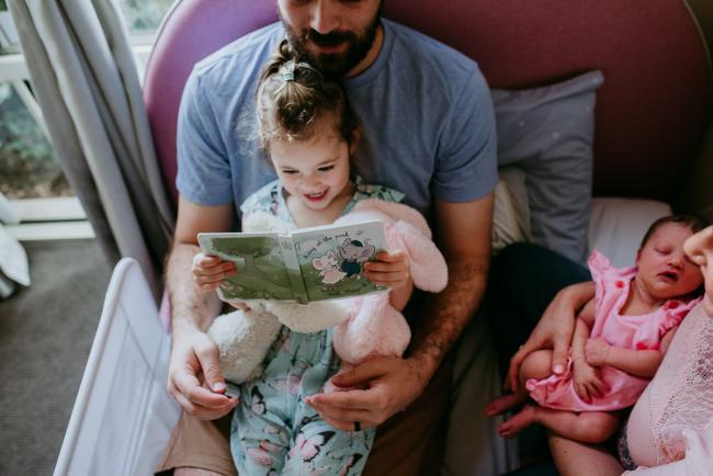 Little girl sits on her dad's lap as they read a story during a Perth newborn lifestyle photography session