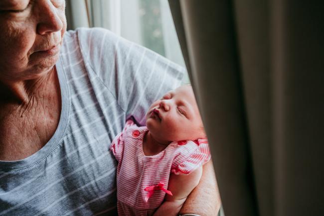 Grandmother holding her new baby granddaughter by a window and looking down on her during a Perth newborn lifestyle photography session