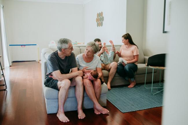 Grandparents sitting on a couch and smiling as they hold their new baby granddaughter as the parents and older sibling sit and play behind them during a Perth newborn lifestyle photography session