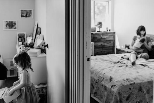 Black and white image of mother holding her new baby on her bed in one room, and their toddler daughter playing in another room during a newborn lifestyle photography Perth session