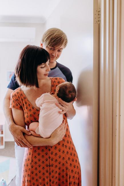 Mother and father stand in the doorway as they look down at their new baby who the mum is holding during a Perth lifestyle Newborn photography session