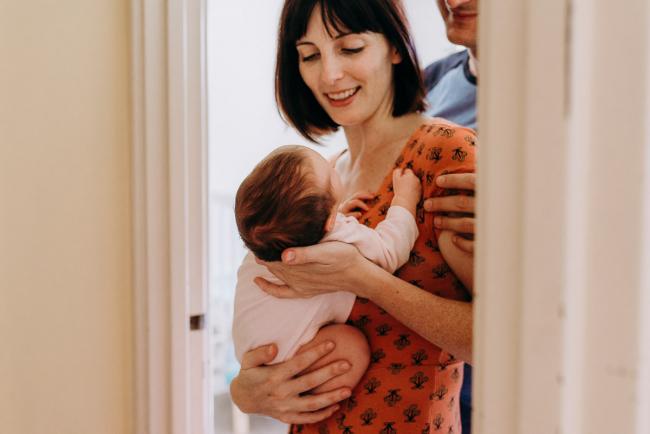 Side image of a mother walking around the corner holding her new baby and smiling as her husband stands behind her during a Perth lifestyle Newborn photography session