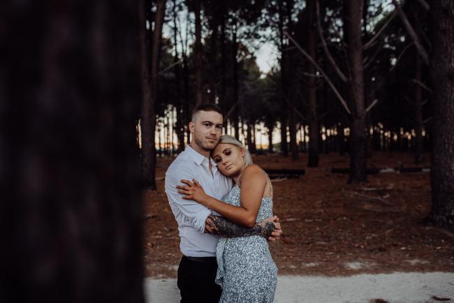 Perth-Couples-Photography-20-of-27 Couple embracing with the man looking at the camera while the woman leans against his chest at The Pines Wanneroo during a Golden Hour Perth couples photography session