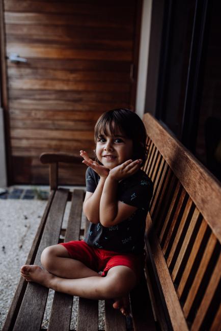 Perth-Family-Photographer-14-of-26 Little boy smiling and framing his face with his hands as he sits on a wooden chair with family photographer Perth