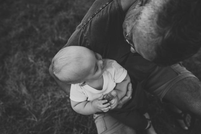 Black and white top down image of baby grandson sitting on his grandfather's lap during a Perth extended family photography session