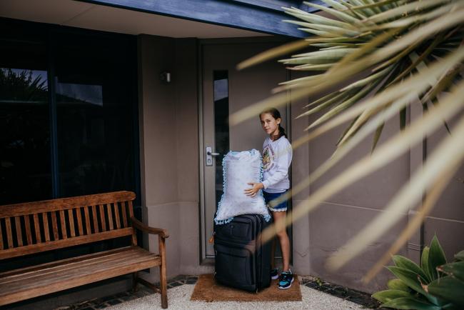 Perth-Family-Photographer-2-of-26 Girl standing outside with her pillow and a suitcase with the image taken through a tree with Perth family photographer