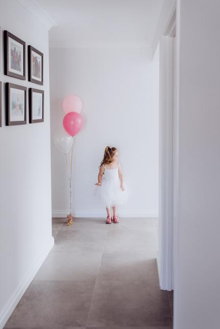 Little girl standing at the end of the hallway next to two pink balloons during a birthday party captured by Perth events photographer