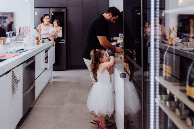 Little girl reaching up to take food her father is preparing in the kitchen during a birthday party captured by Perth events photographer