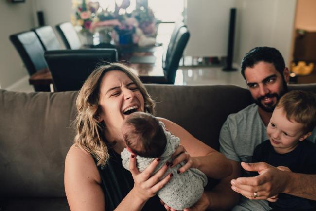 Mother laughing as her new baby is being passed to her on the couch with dad and brother smiling during a Perth lifestyle newborn photography session