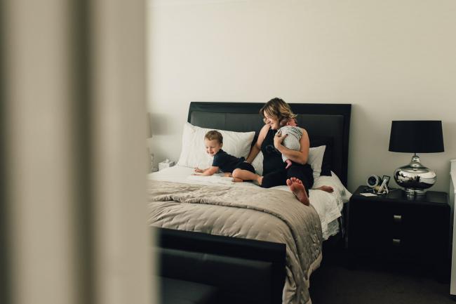 Looking through the doorway as a mother sits on the bed holding her new baby, and next to her toddler during a Perth lifestyle newborn photography session