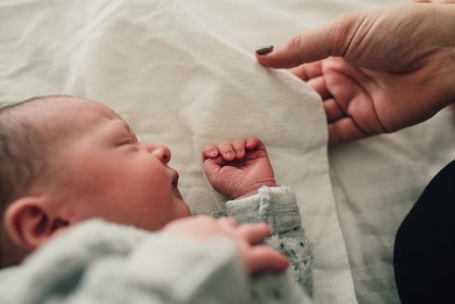 New baby laying on a bed with her face towards her hand, and her mother's hand coming into the frame during a Perth lifestyle newborn photography session