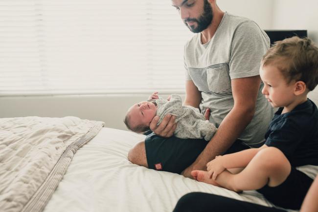 Father holding new baby out on the bed next to brother during a Perth lifestyle newborn photography session