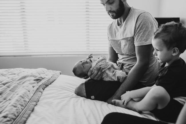 Black and white image of father holding new baby out on the bed next to brother during a Perth lifestyle newborn photography session