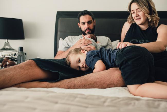 Little boy laying across the legs of his father as dad holds the new baby on the bed next to mum during a Perth lifestyle newborn photography session