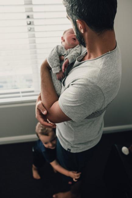 Father holding his new baby while his son wraps his arms around his legs during a Perth lifestyle newborn photography session