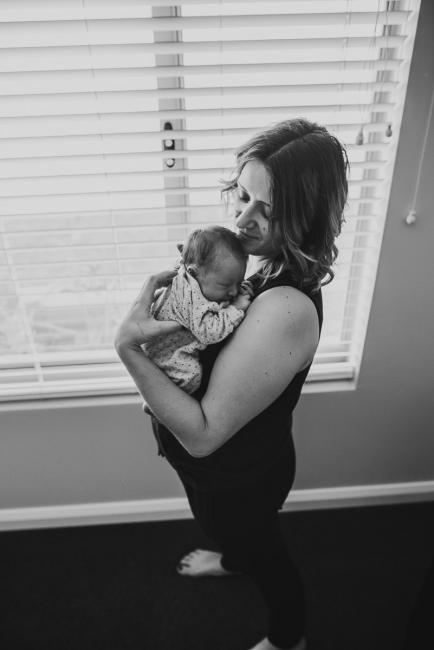 Black and white image of mother holding new baby next to a window during a lifestyle newborn photography Perth session