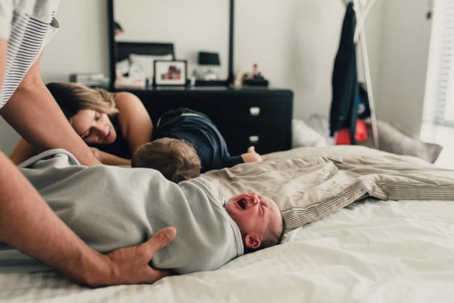 Wrapped baby on a bed crying as dad lifts her and mum and brother lay with thier heads on the bed during a Perth lifestyle newborn photography session