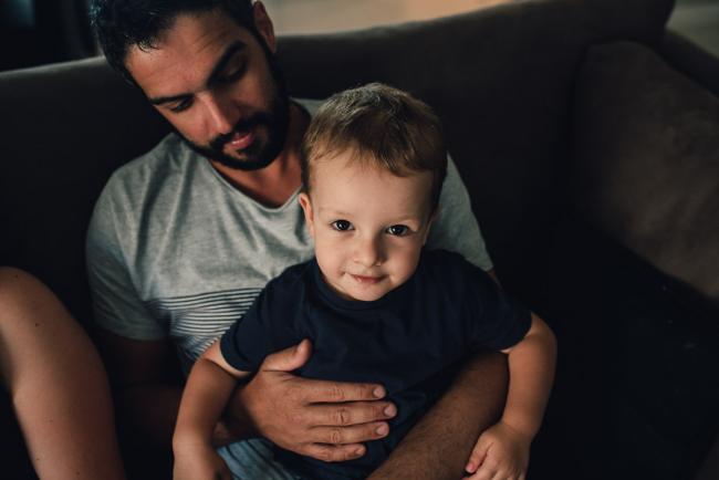 Little boy sitting on his dad's lap and looking up at the camera during a Perth lifestyle newborn photography session
