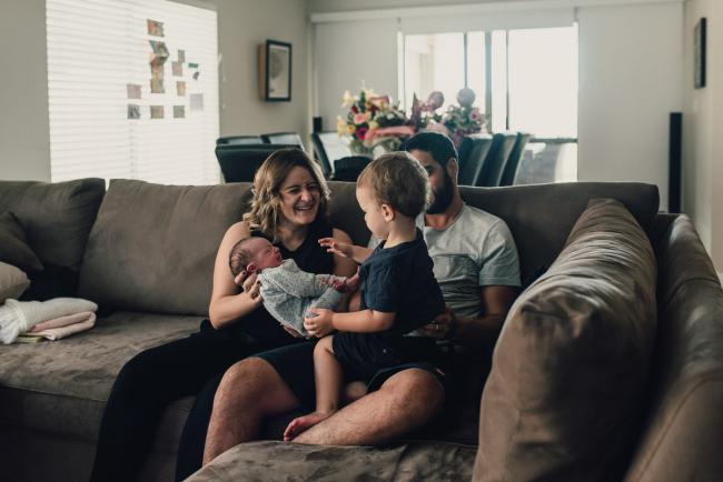 Little boy sitting on his dad's lap and being passed his new baby sister by his mother during a Perth lifestyle newborn photography session