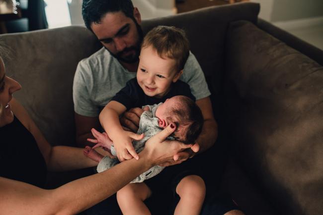 Little boy smiling as he's being passed his new baby sister to hold whilst sitting on his dad's lap during a Perth lifestyle newborn photography session