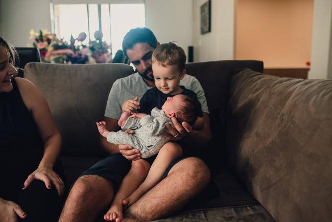 Brother sitting on his dad's lap as he holds his new baby sister and mum looks on during a Perth lifestyle newborn photography session