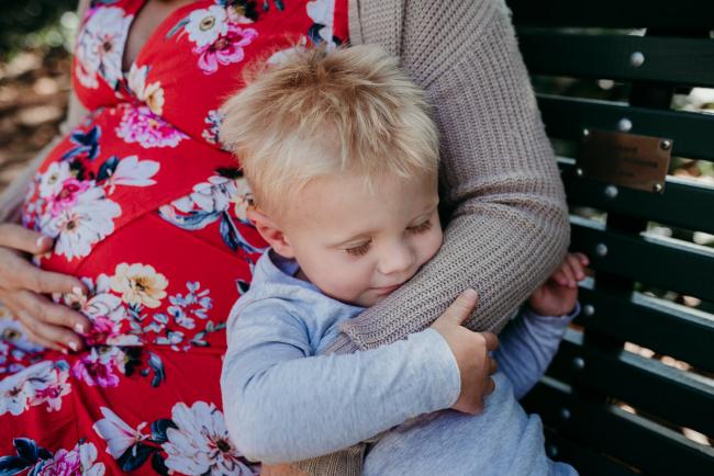 Little boy sitting on a bench next to his pregnant mother and cuddling her arm during a Perth maternity session at Hyde Park