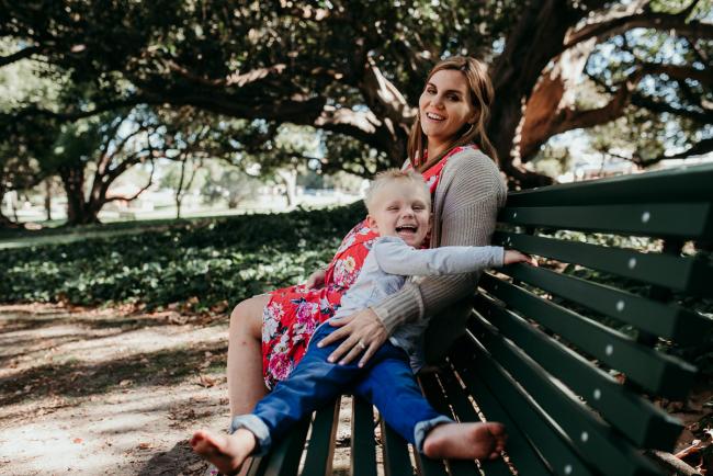 Little boy laughing at the camera as he sits on a green bench next to his pregnant mother during a Perth maternity session at Hyde Park