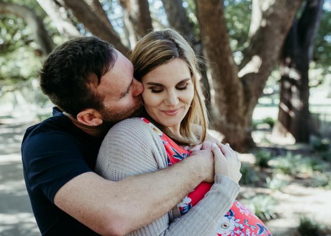 Husband hugging his pregnant wife from behind as she smiles and looks down during a Perth maternity session at Hyde Park
