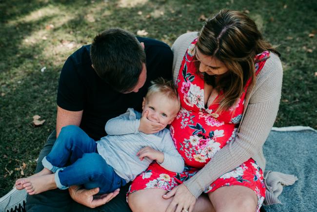Little boy lying across the lap of his parents as he covers his mouth during a Perth maternity session at Hyde Park