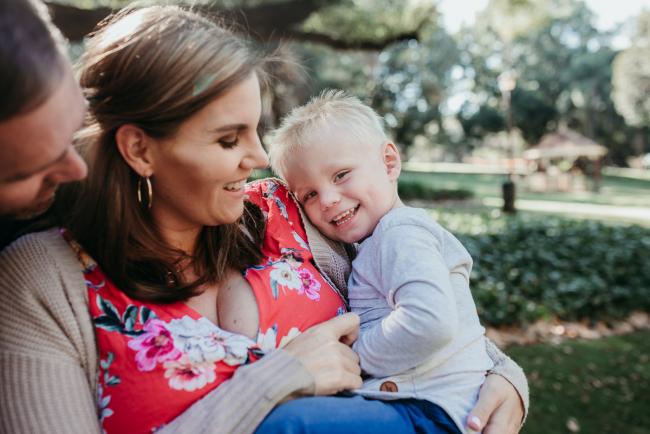 Little boy laughing at the camera as his mother holds him and dad looks in from the side during a Perth maternity session at Hyde Park