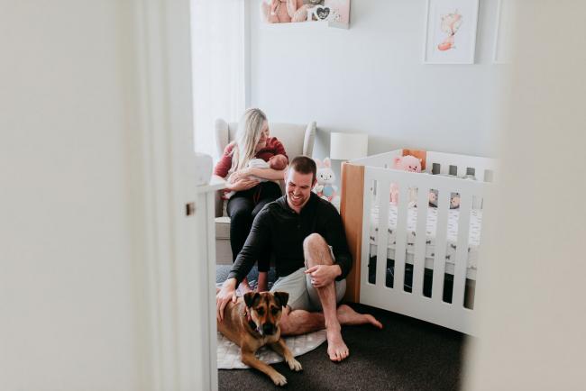 Newborn-Photographer-Perth-13-of-37 Mother sitting with a newborn baby in the chair while the father and dog sit on the floor by their feet during a Perth newborn lifestyle session