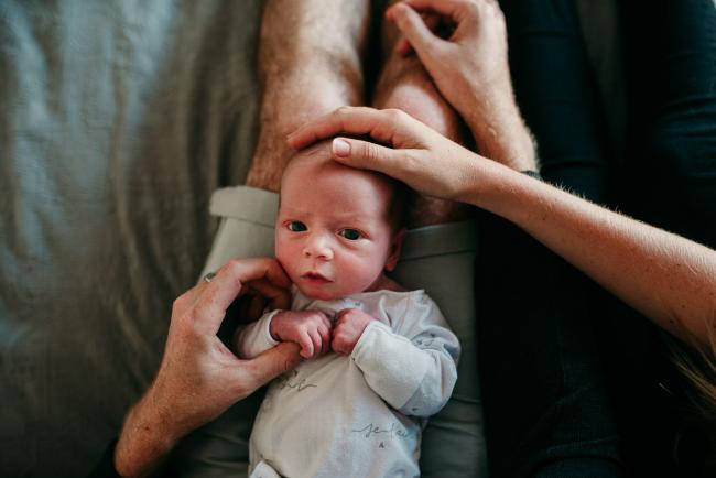 Newborn baby laying on her dad's lap as mum and dad's hands are touching her during a Perth newborn lifestyle session