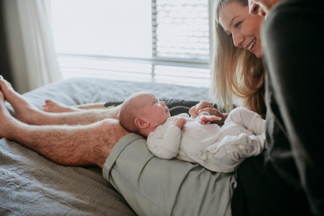 Mum and dad sitting on their bed with their newborn baby laying across dad's lap during a Perth newborn lifestyle session