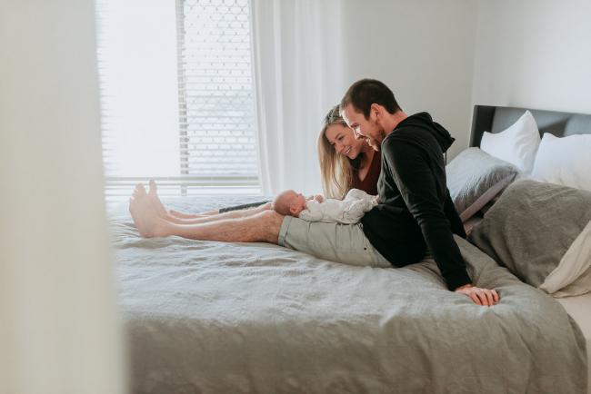 Newborn-Photographer-Perth-18-of-37 Mother and father sitting on the bed with their newborn baby on their lap during a Perth newborn lifestyle session