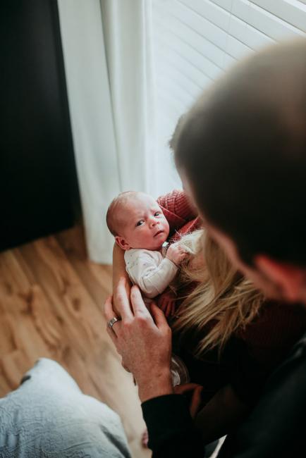 Newborn-Photographer-Perth-2-of-37 Top down image of a newborn baby being held by her mother as her father leans over and touches their arms with Perth lifestyle newborn photographer