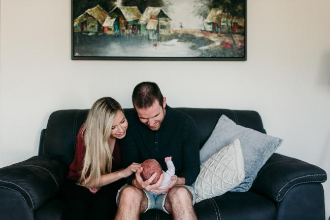 Newborn-Photographer-Perth-22-of-37 Mum and dad sitting on the couch and smiling at their newborn baby during a Perth newborn lifestyle session