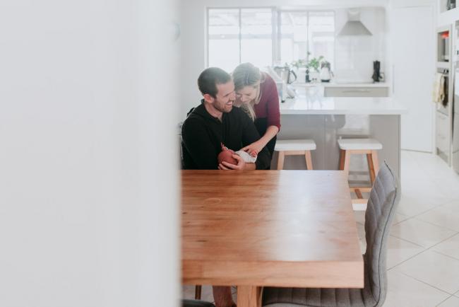 Newborn-Photographer-Perth-25-of-37 Mother and father sitting at a dining table with their newborn baby during a Perth newborn lifestyle session