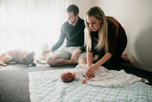 Newborn-Photographer-Perth-26-of-37 Mother wrapping her newborn baby on the floor as dad sits behind with the dog during a Perth newborn lifestyle session
