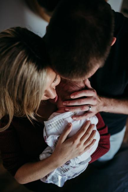Newborn-Photographer-Perth-33-of-37 Top down image of mother holding her newborn baby on her chest as dad holds and kisses the baby during a Perth newborn lifestyle session
