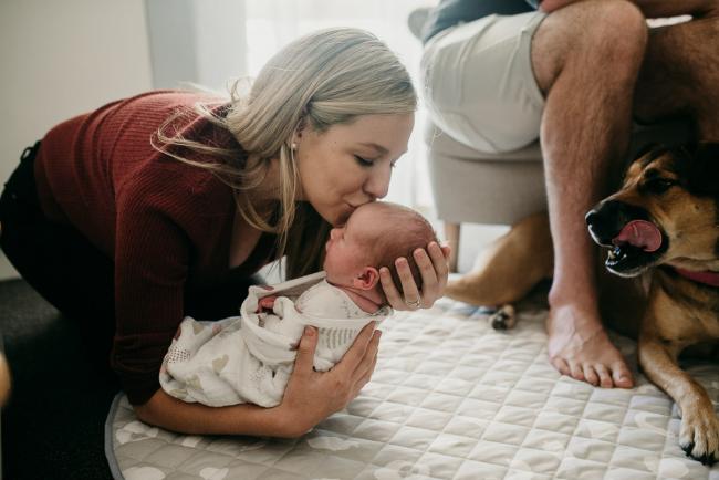 Newborn-Photographer-Perth-36-of-37 Mother lifting and kissing her newborn baby with the dog and dad's feet in the frame during a Perth newborn lifestyle session