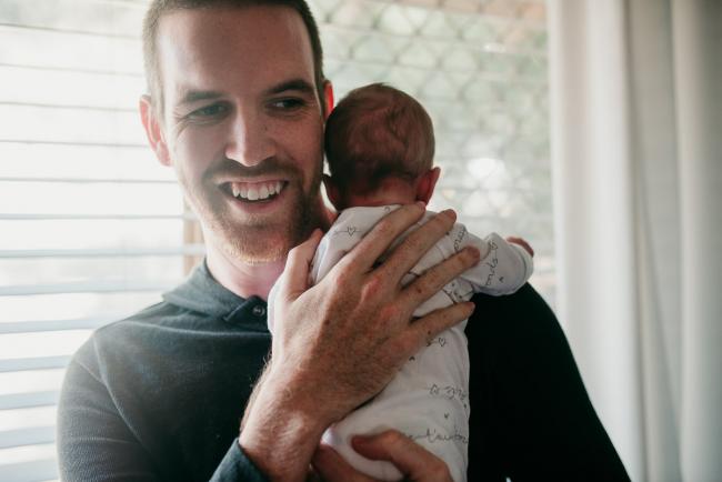 Newborn-Photographer-Perth-6-of-37 Newborn baby being held over her fathers shoulder as he smiles during a Perth newborn lifestyle session
