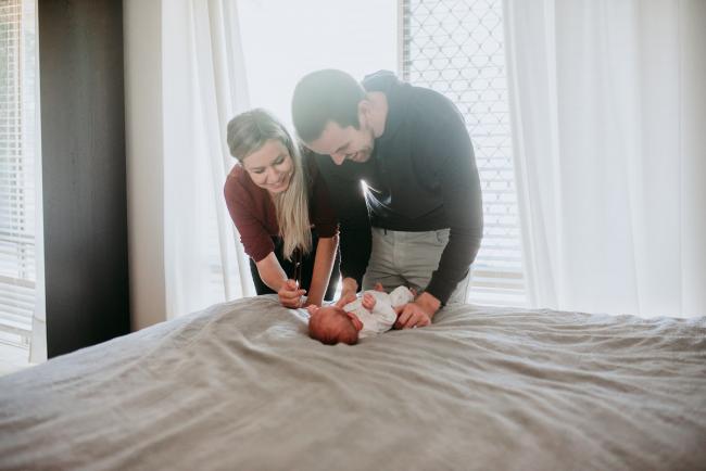 Newborn-Photographer-Perth-8-of-37 New parents smiling down at their newborn baby who is laying on the bed during a Perth newborn lifestyle session