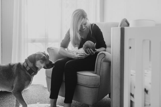 Newborn-Photographer-Perth-9-of-37 black and white image of mother sitting in a chair in the nursery with her newborn baby as the dog comes into the frame during a Perth newborn lifestyle session