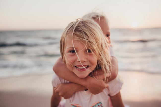 Little girl smiling at the camera as she gives her little sister a piggyback at Burns Beach during a Perth family photography session