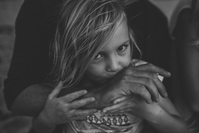 Black and white image of little girl holding her dad's hand and kissing it as she looks at the camera at Burns Beach during a Perth family photography session