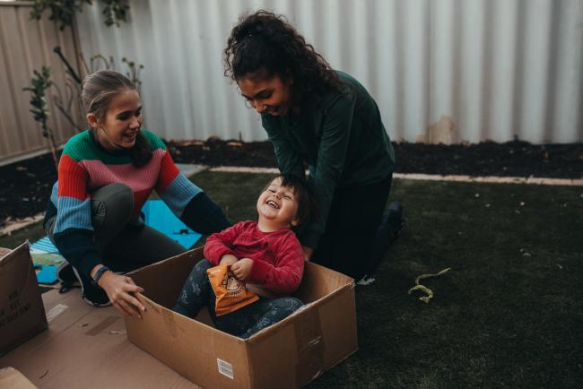 20190422-DSC_2949 Little boy laughing as he sits in a box with two older girls holding him with Perth family photographer